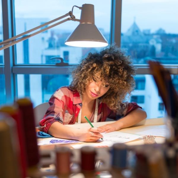 Designer sidder ved skrivebordet under lampen og tegner mode- eller tæppeudkast i et studie.